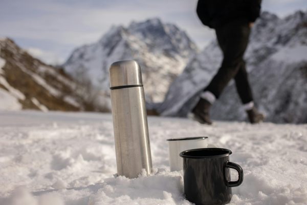 A person hikes in snowy mountains with a thermos and mugs on the snow, showing winter adventure.